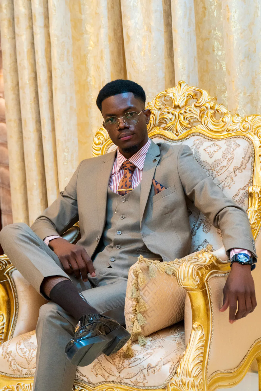 Kayesi Blessed seated in a gilded chair, wearing a three-piece suit